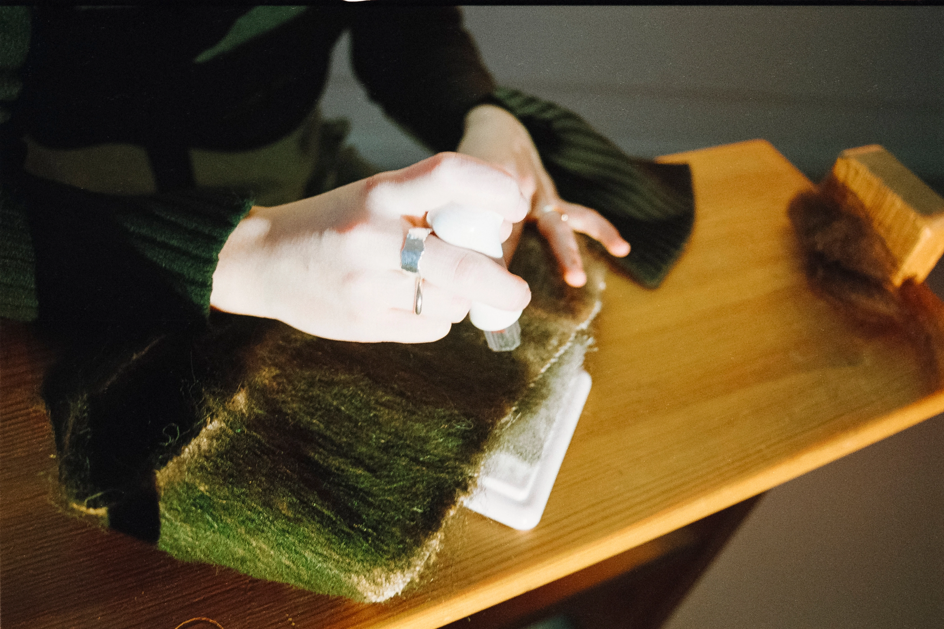Analog photograph of a hand dyeing wool fibers green on a table, with a small bottle of dye.